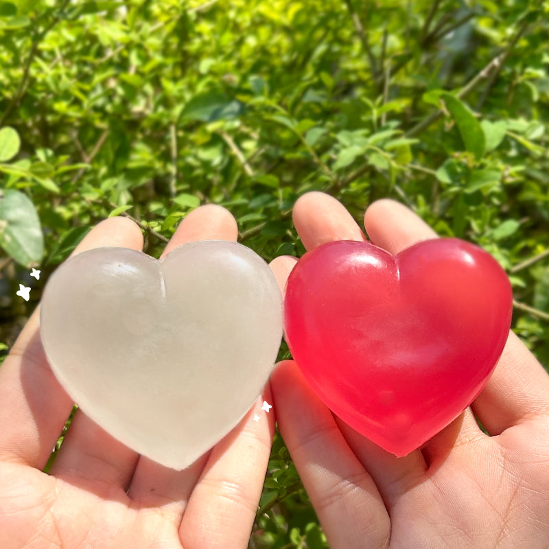 Two heart-shaped objects, one clear and one red, held in hands with a green leafy background.